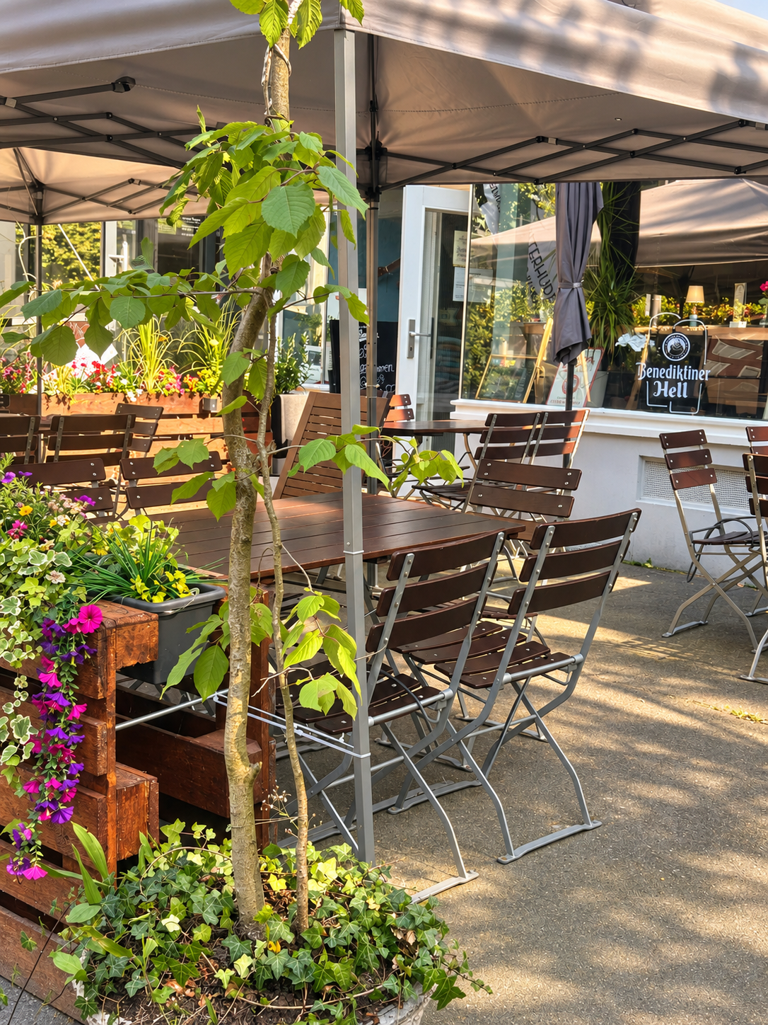 Clax-Terrasse am Tag: Paletten-Beet mit Blumen, Tische und Stühle unter grauem Sonnenschirm